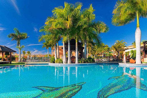 A luxurious and sunny resort pool, palm trees and scattered sheltered sun bed huts. The pool depicts dolphins created using coloured mosaic tiles forming the foundations of the pool