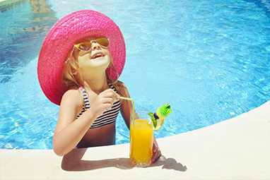 Young girl sipping orange cocktail in a pool with a pink hat and swimming costume